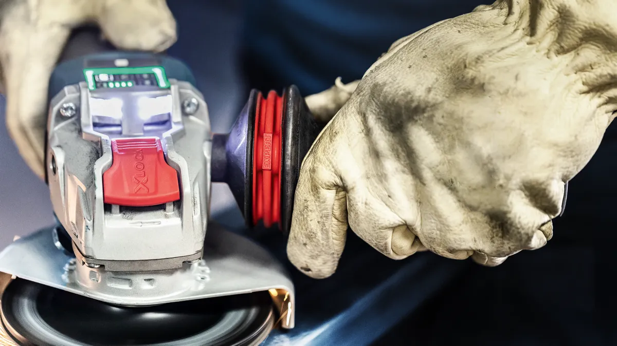 Oseba, ki nosi varnostno opremo, upravlja kotni brusilnik na kovinski površini.